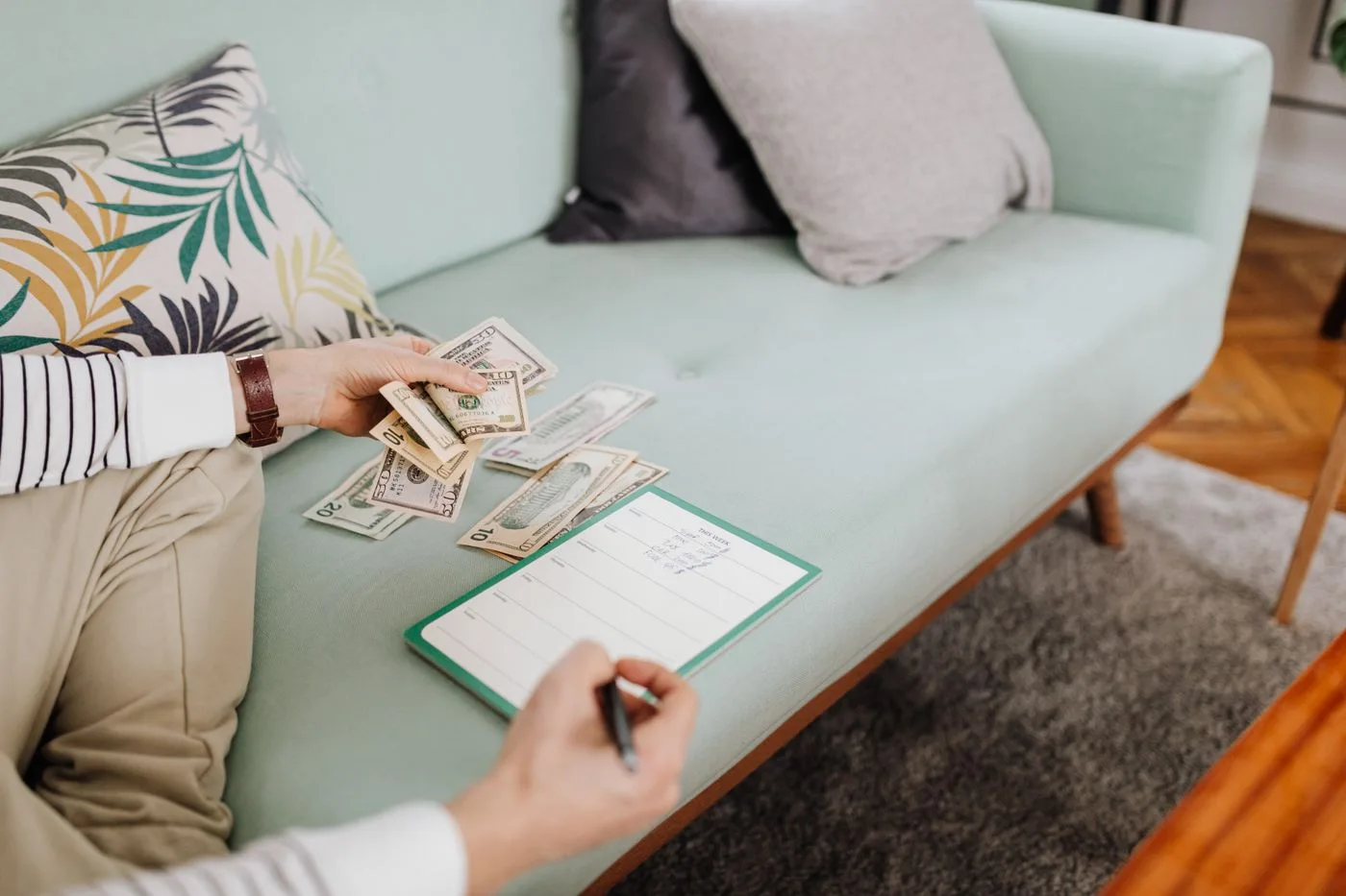 Adult counting cash and writing in a notebook while seated on a stylish couch at home.