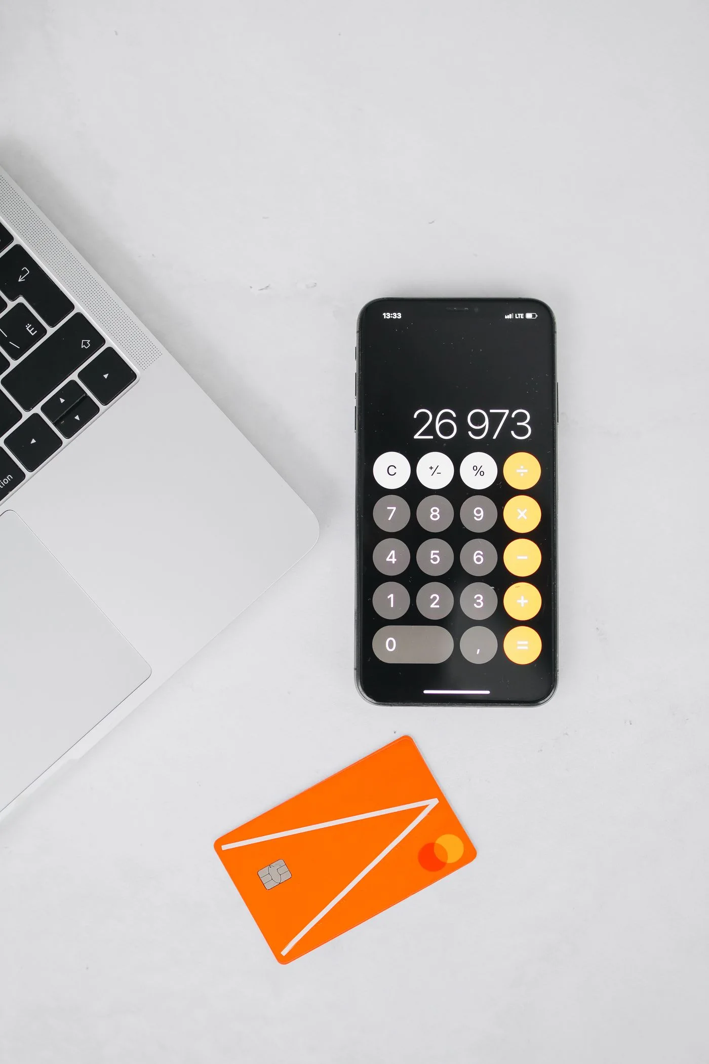 A flat lay of a laptop, smartphone with calculator app, and orange card on a white surface.