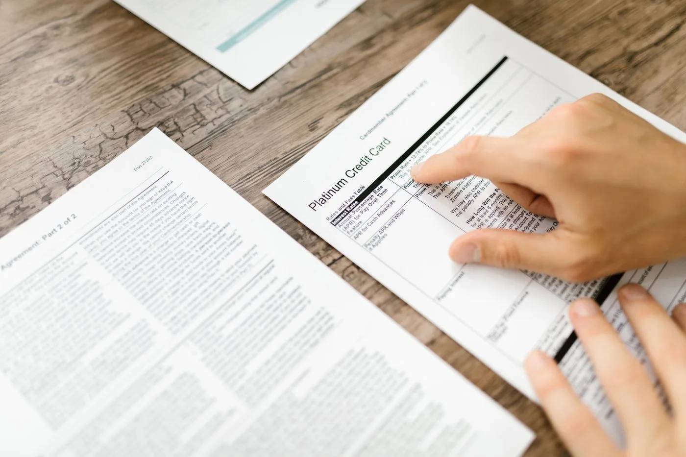A hand examining a credit card agreement on a wooden desk, highlighting financial review.